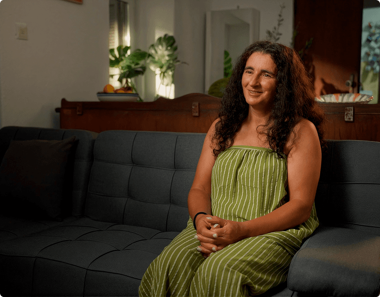 A woman with long dark curly hair wearing a green and white striped jumpsuit sits on a dark gray couch.