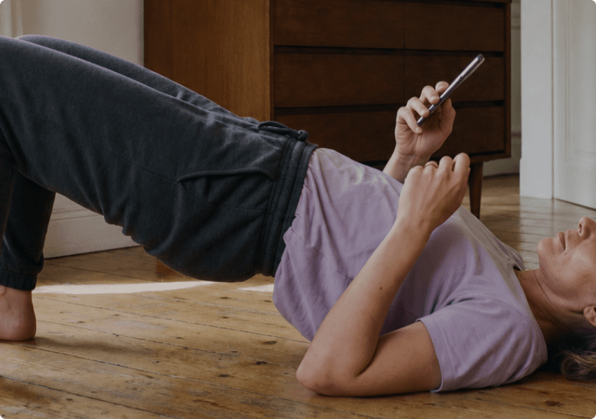 Person lying on a wooden floor, holding a smartphone while their legs are bent, wearing a purple shirt and dark sweatpants.