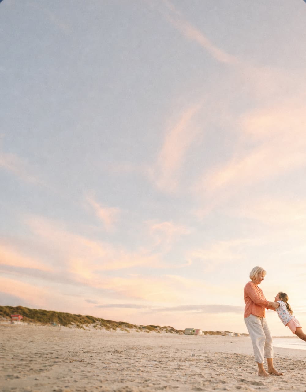 A woman and a child play on a sandy beach at sunset, with soft pink and orange clouds in the sky.