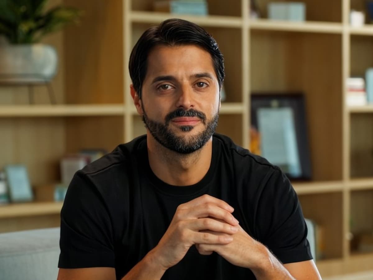 A man with short dark hair and a beard, wearing a black shirt, sits indoors with hands clasped, in front of a shelf with books and decor.