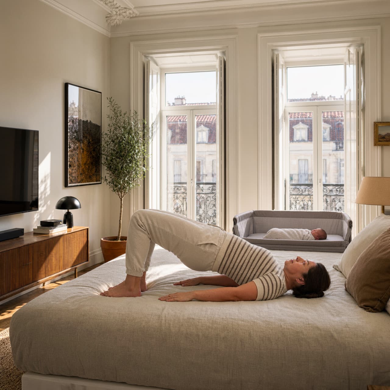 A woman performs a glute bridge exercise on a bed while a baby sleeps in a bedside bassinet nearby.