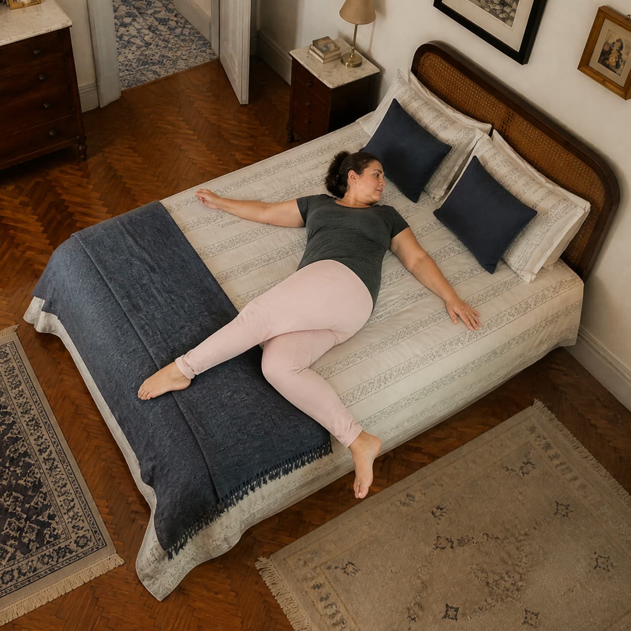 A person in a gray t-shirt and pink pants lies spread-eagle on a bed with a wooden headboard, dark blue pillows, and a matching throw blanket.