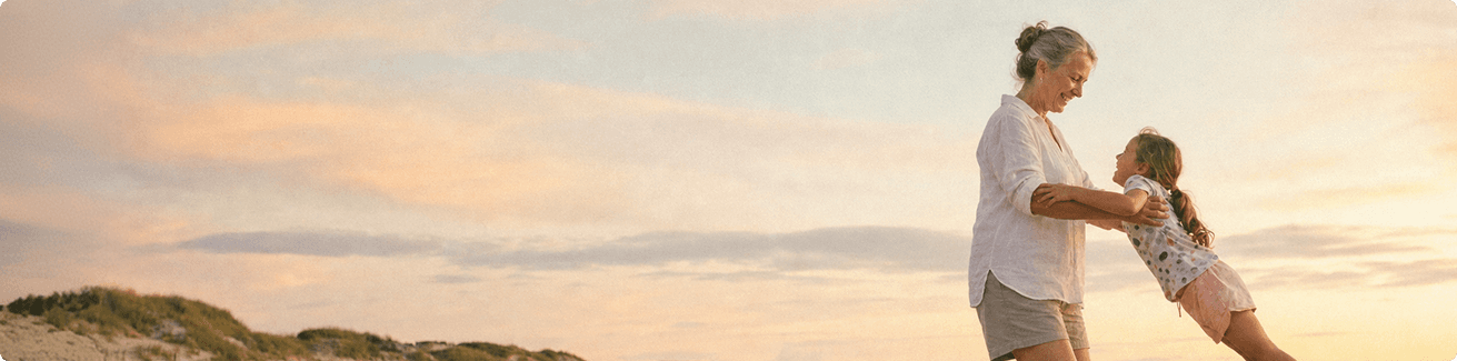 A woman joyfully swings a young girl by the arms on a beach at sunset, with sandy dunes and a pastel sky in the background.