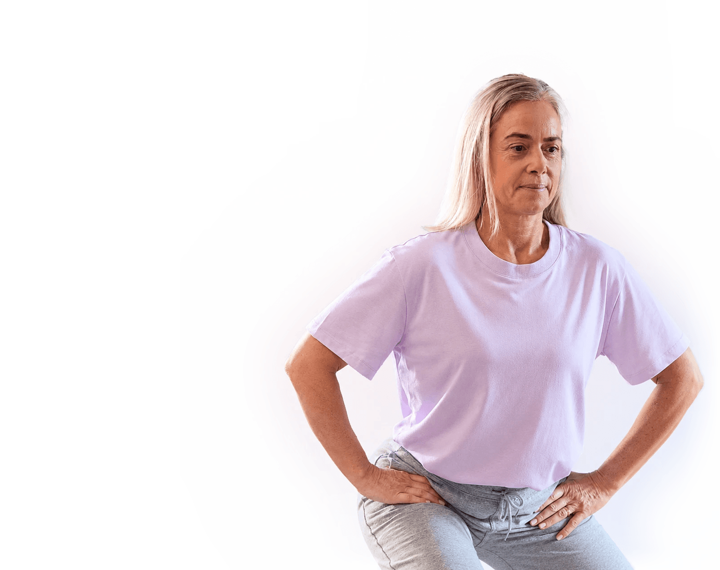 A woman with long blond hair wearing a lavender T-shirt and gray pants is exercising, posing with her hands on her hips against a white background.