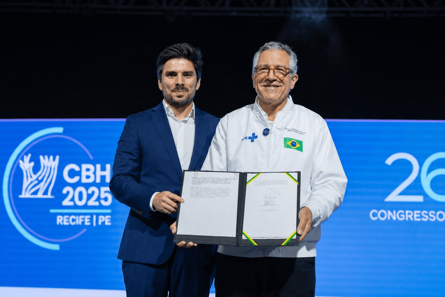 Two men hold a document on stage at an event. The backdrop reads "CBH 2025" with a logo and "Recife | PE".