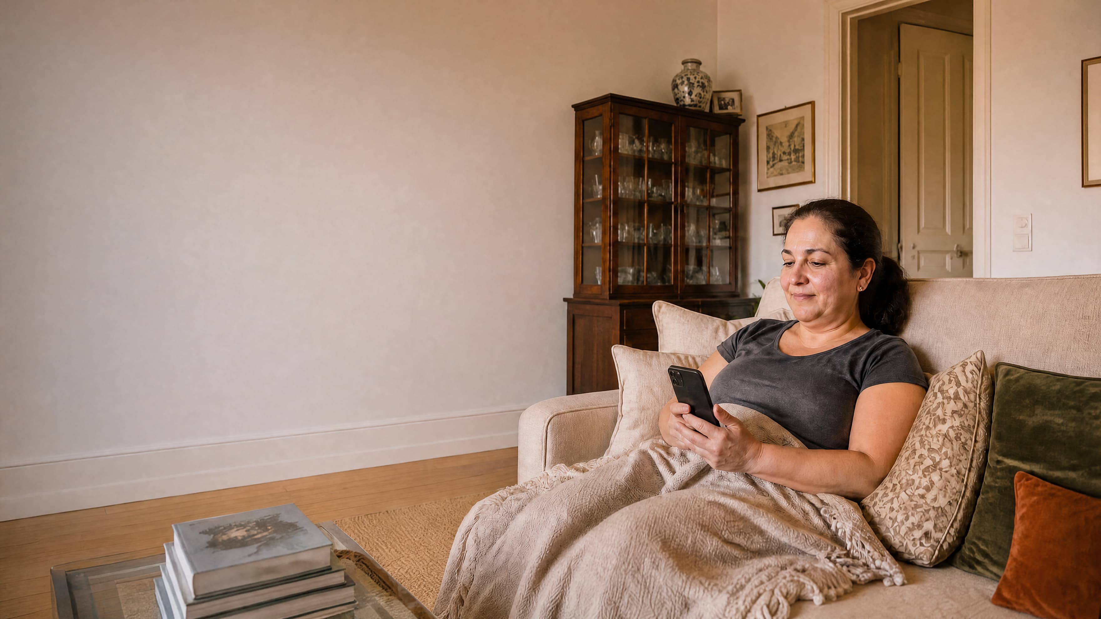 A woman sits on a sofa covered by a blanket, looking at her smartphone.