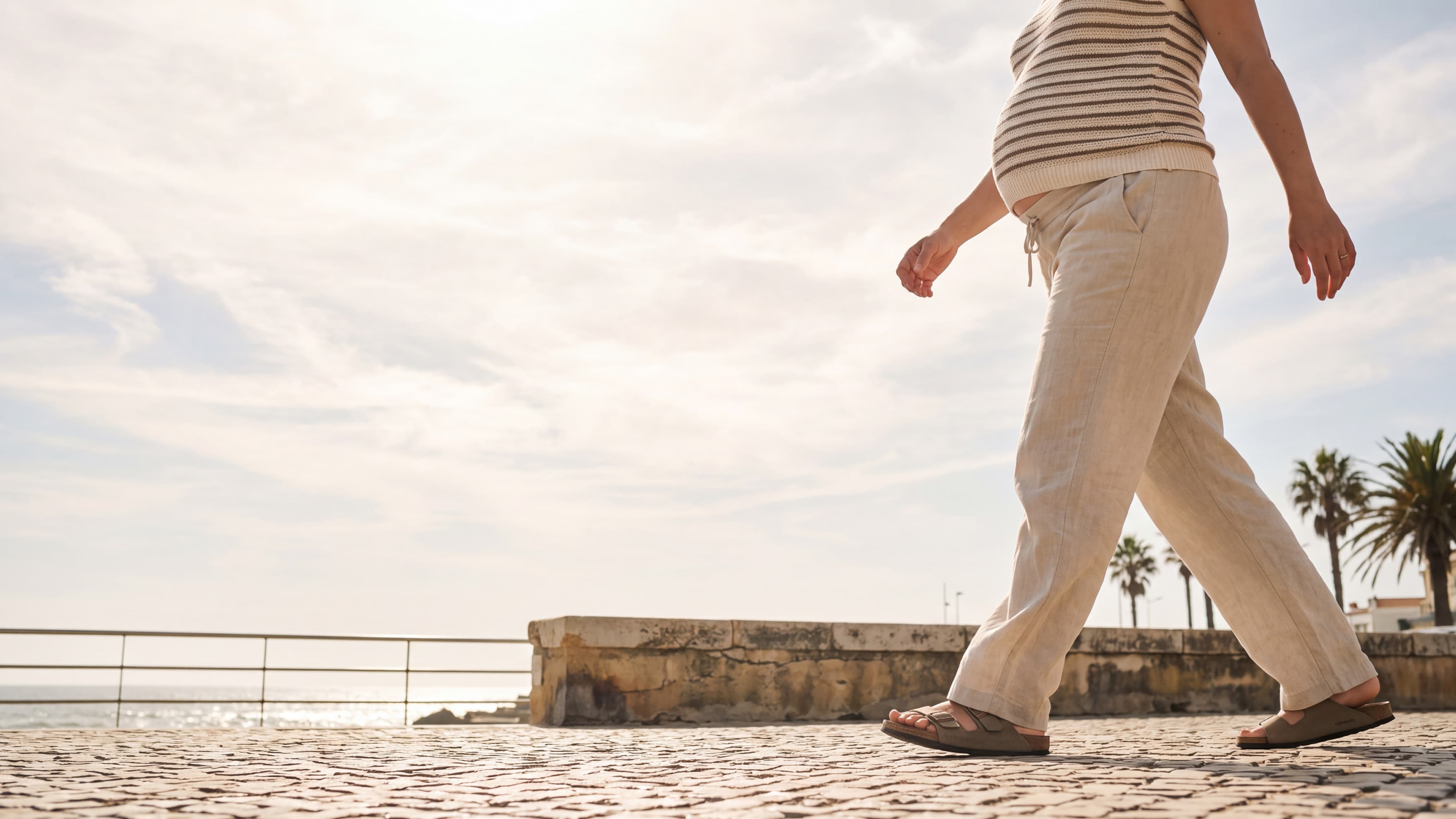A pregnant person in a striped top and light-colored trousers walking along a coastal promenade.
