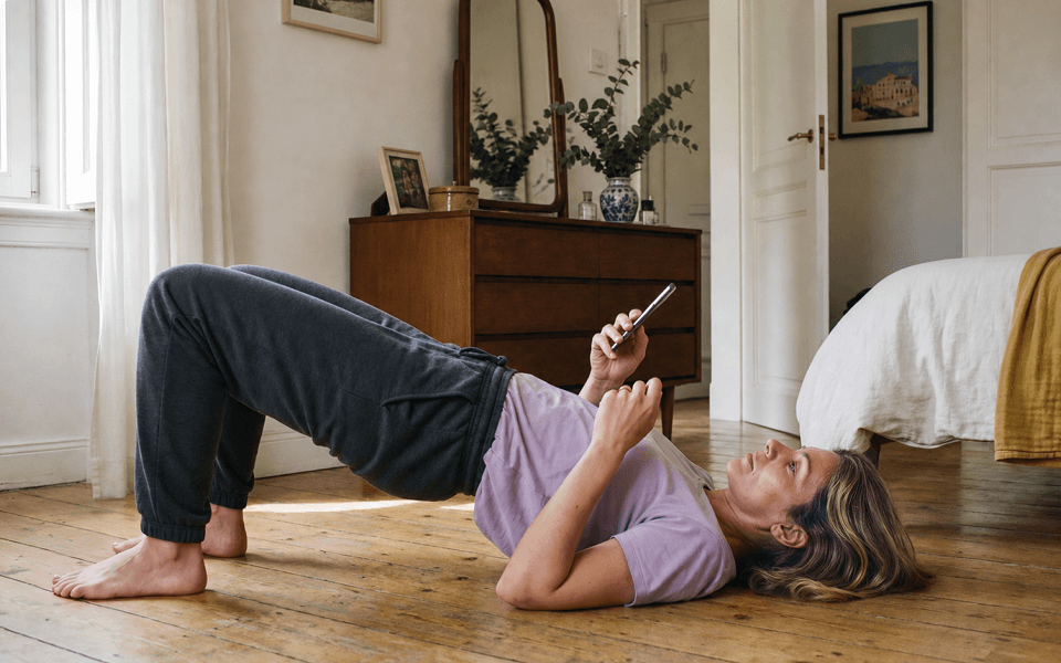 Woman lying on the floor in a bridge position, holding a phone, in a cozy bedroom with a wooden dresser and decor.